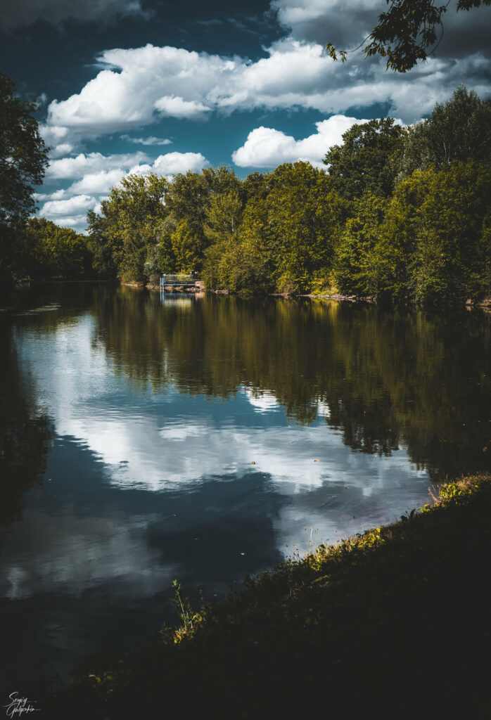 Tranquil river with lush green trees and wooden pier reflected in calm water under blue sky Wallpaper