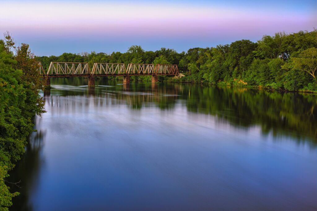 Tranquil river with reflective water lush green trees rustic bridge under lilac blue sky wallpaper