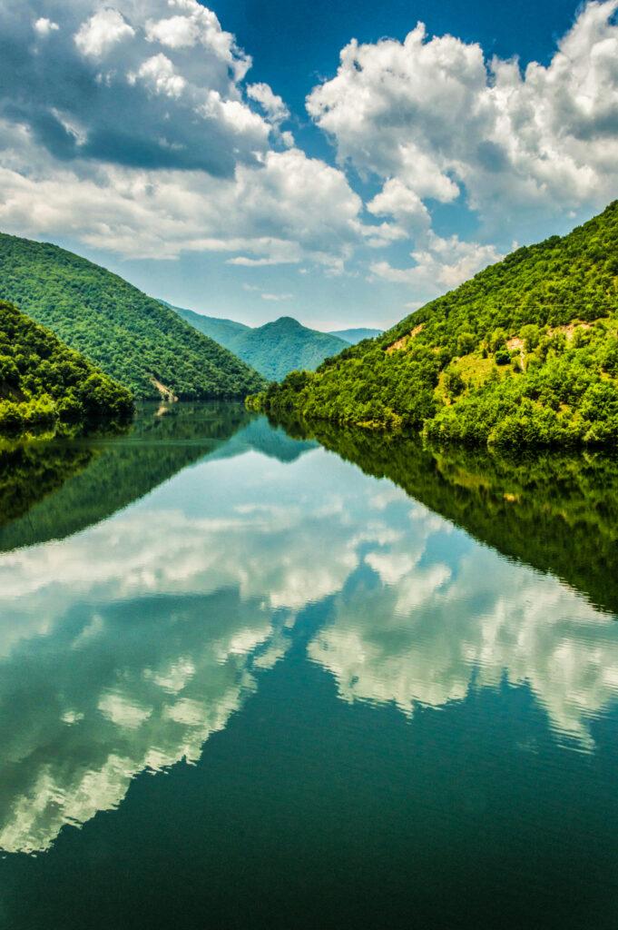 Tranquil river flowing through lush green forested hills reflecting vibrant blue sky clouds Wallpaper