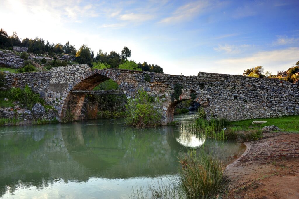 Tranquil river flowing under ancient stone bridge surrounded by lush greenery and rugged landscape Wallpaper
