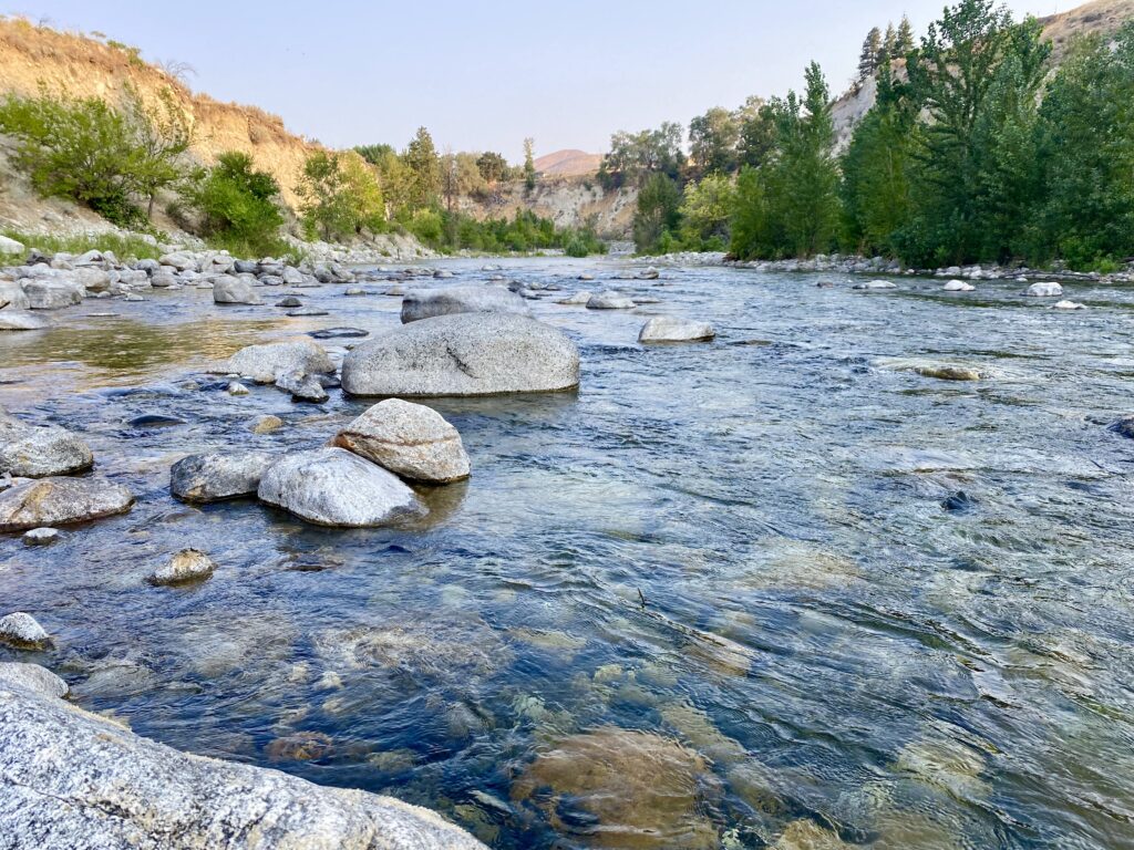 Serene river flowing over smooth stones with green trees and hills in a peaceful nature scene Wallpaper
