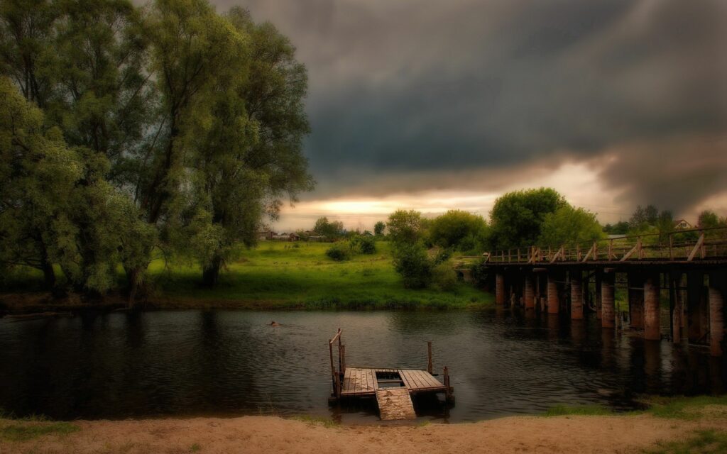 Tranquil riverside sunset with wooden dock lush grass rustic bridge and dramatic cloudy sky Wallpaper