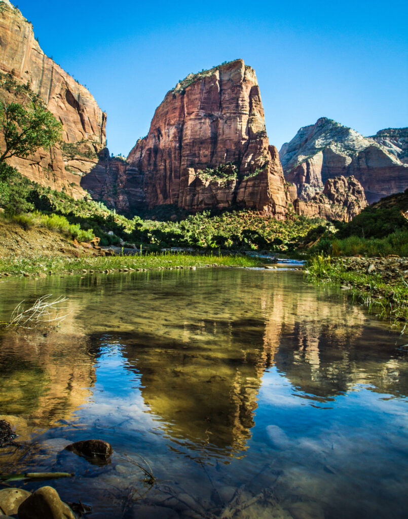 Towering rocky cliffs reflected in calm pond with lush greenery and vibrant blue sky background Wallpaper