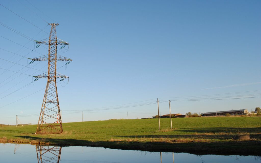 Tranquil rural landscape with calm river green field and towering electrical pylon under blue sky Wallpaper