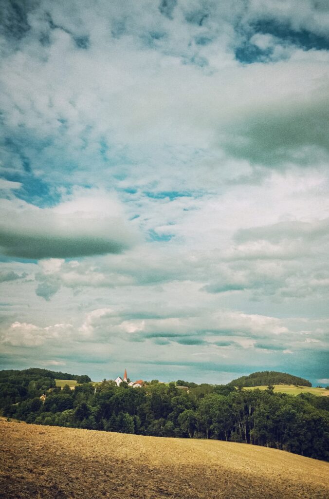 Tranquil rural landscape with rolling fields dense forest quaint buildings and dramatic sky clouds Wallpaper