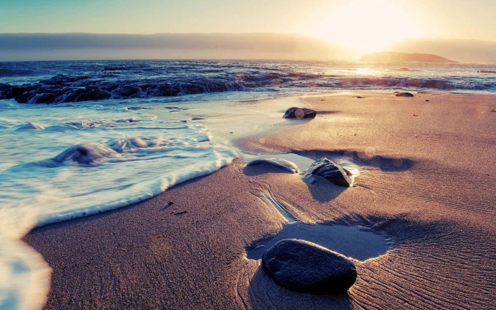 Tranquil beach with wet sand dark rocks and gentle ocean waves at golden sunset wallpaper background