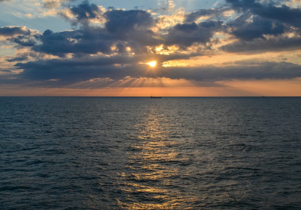 Tranquil sea with gentle waves under dramatic sunset sky sunbeams on distant ship wallpaper