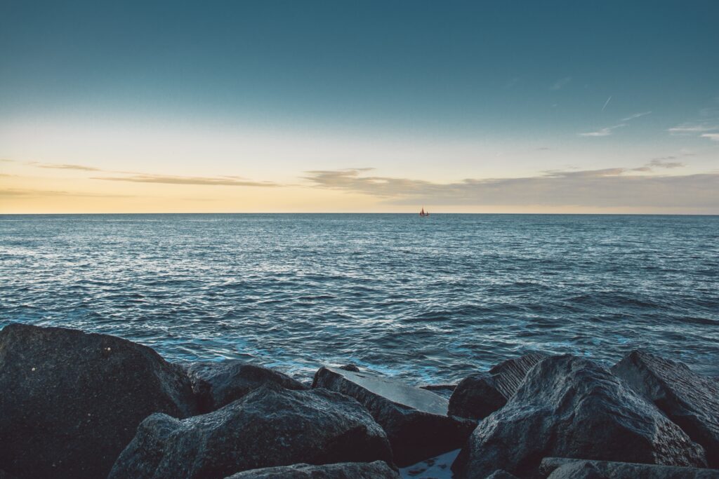 Tranquil sea with gentle waves and dark stones under golden sky with distant boat wallpaper