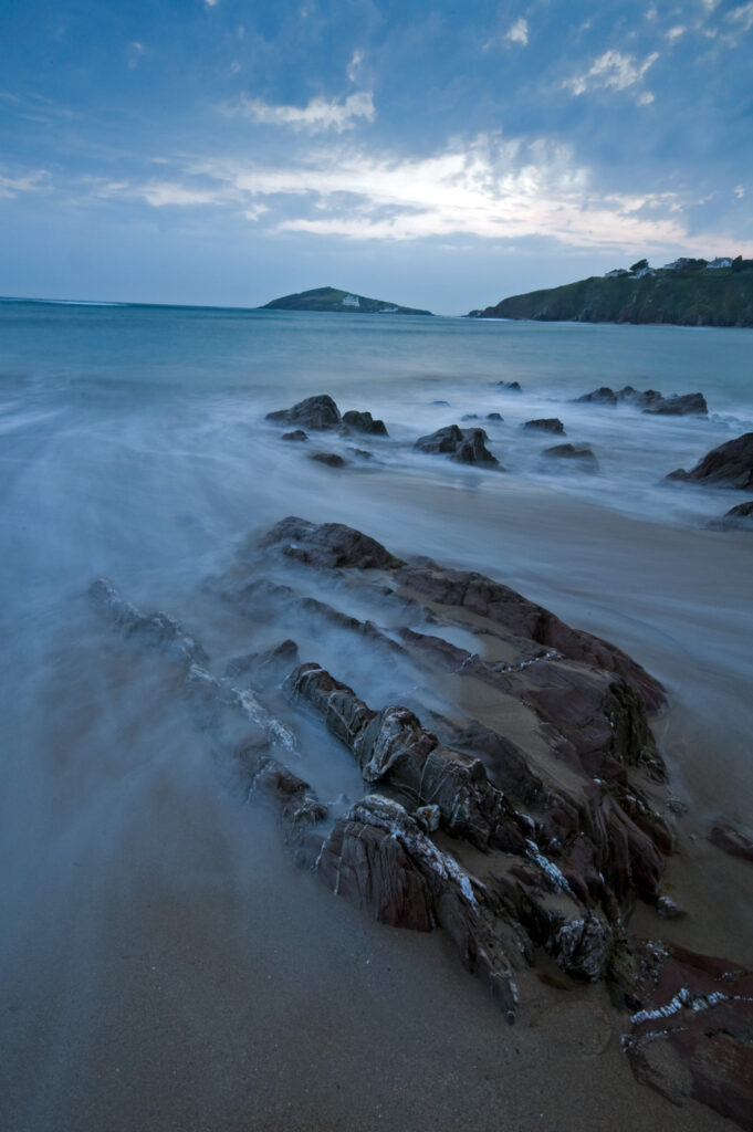Tranquil seascape with smooth waves over rocky shore under soft cloudy blue sky wallpaper background