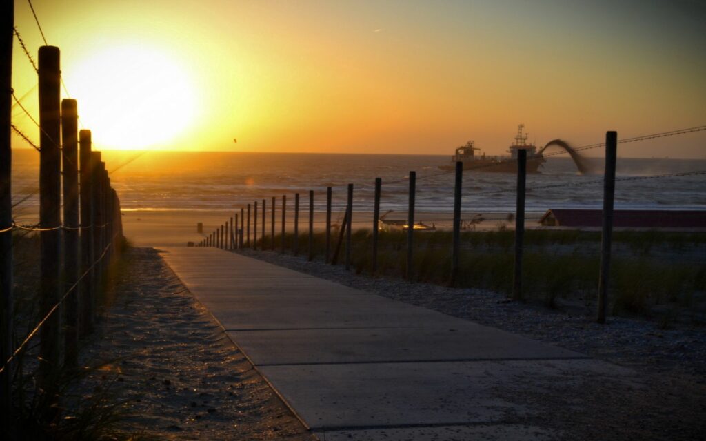 Tranquil seaside sunset with paved path sandy beach calm waves golden light dune grasses Wallpaper