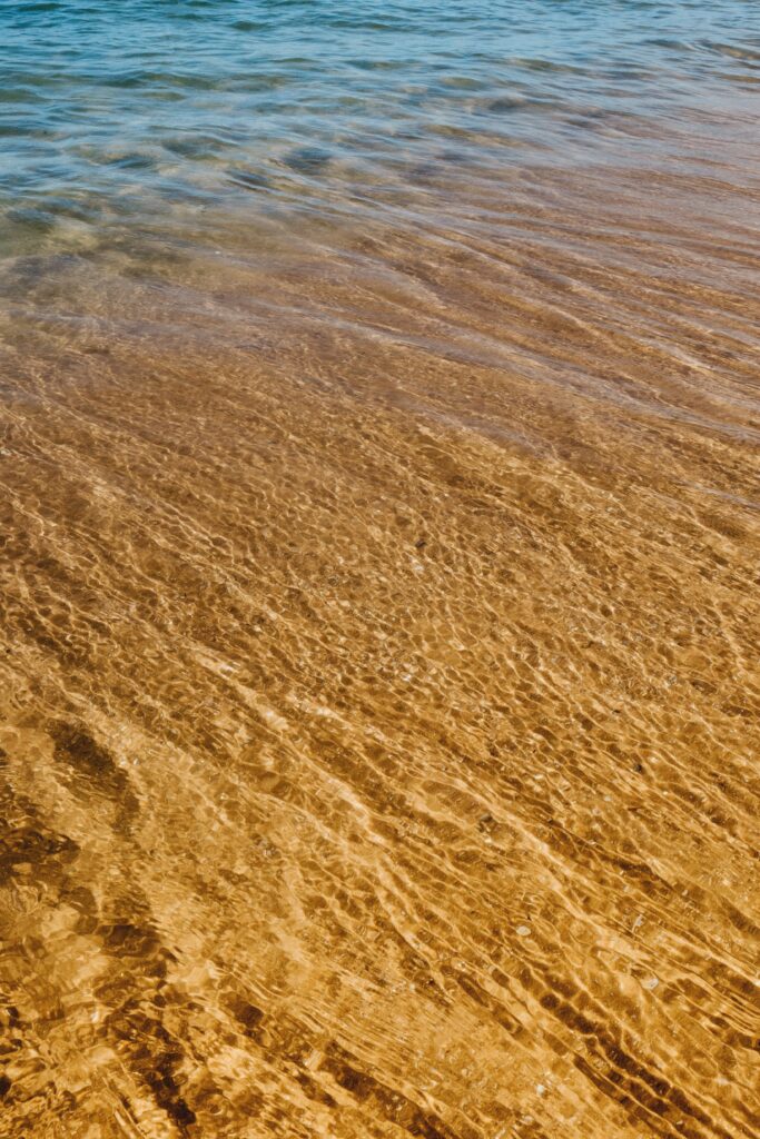 Transparent water ripples over golden sand with sunlight highlighting serene shallow shoreline patterns Wallpaper