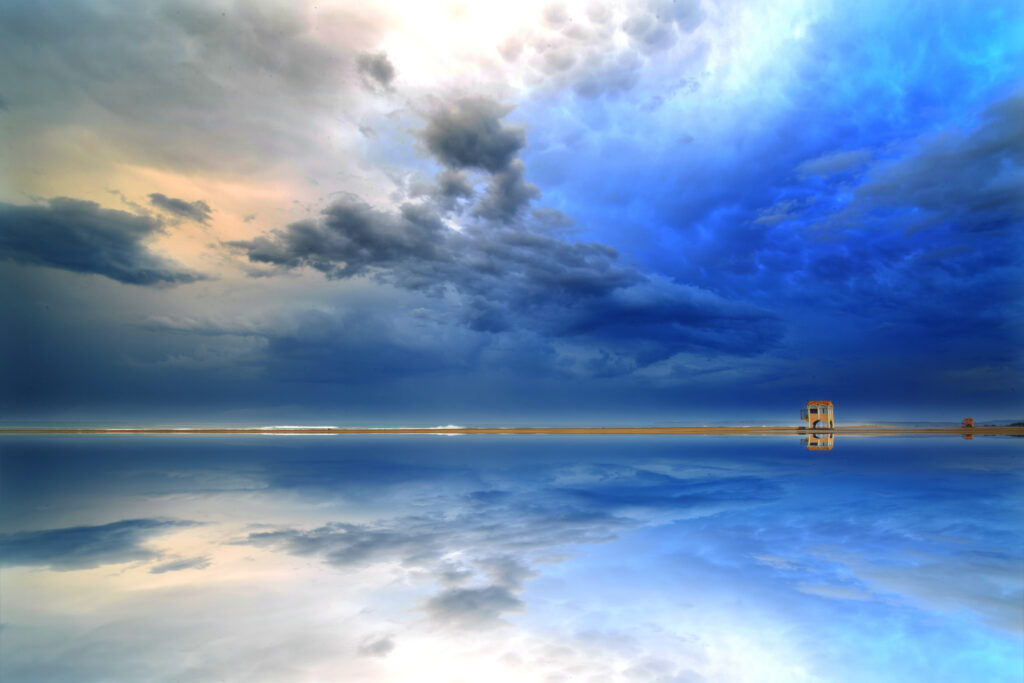 Serene coastal shore with dramatic clouds reflecting on calm water and a distant lifeguard tower Wallpaper