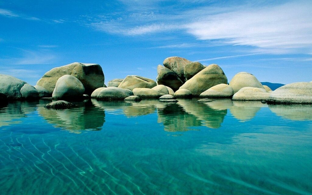 Serene wallpaper with smooth stones reflected on clear blue water ripples and bright sky background