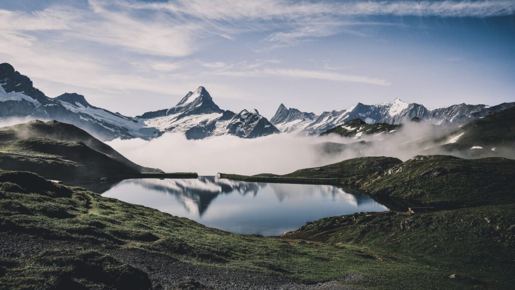 Snow-capped mountains reflected in lake with foggy green hills under clear blue sky wallpaper