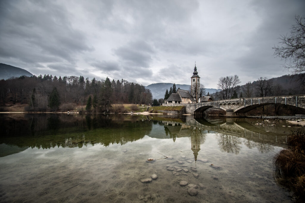 Stone bridge and chapel by calm river with bare trees under cloudy sky serene wallpaper background