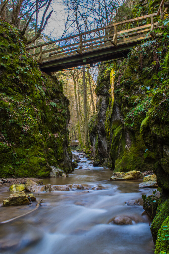 Tranquil stream over smooth stones under rustic wooden bridge with mossy cliffs and sunlight filtering Wallpaper