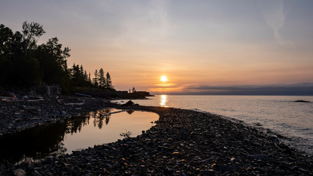 Tranquil sunrise over serene pebble coastline with tidal pools silhouetted trees and pastel sky Wallpaper