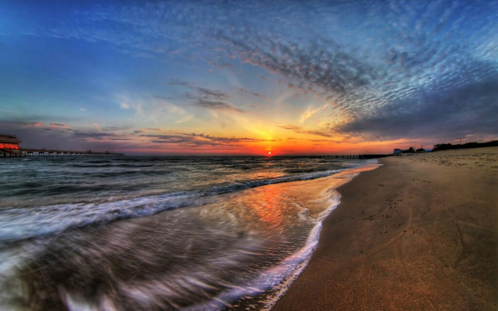 Sunset beach wallpaper with glowing sun reflecting on waves and wet sand under wispy clouds