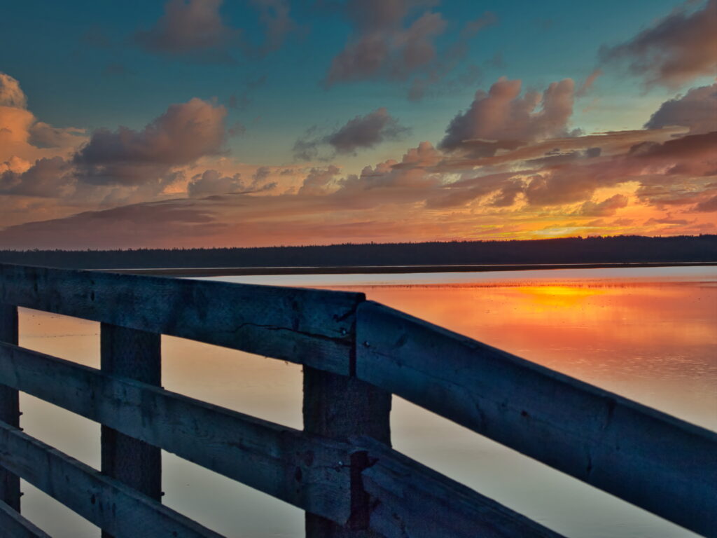 Sunset over tranquil lake with orange blue reflections rustic fence and dramatic clouds background Wallpaper