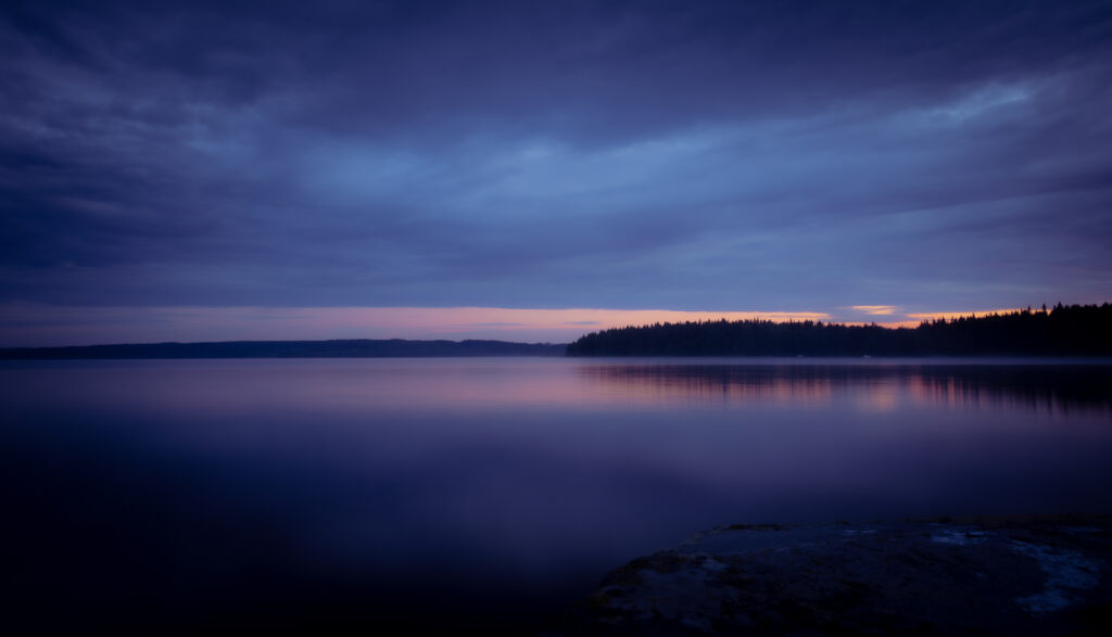 Tranquil sunset lake with calm waters reflecting soft hues and distant tree-lined shore Wallpaper