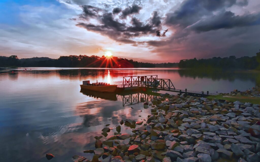 Moored boat at rustic dock on calm lake with sunset reflection stones trees and clouds wallpaper
