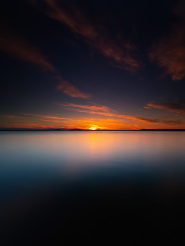 Tranquil sunset over calm waters with glowing sun and twilight sky in deep blues and orange hues Wallpaper