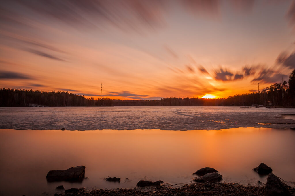 Sunset over icy lake with vibrant orange gold hues smooth stones on shore and blurred clouds Wallpaper