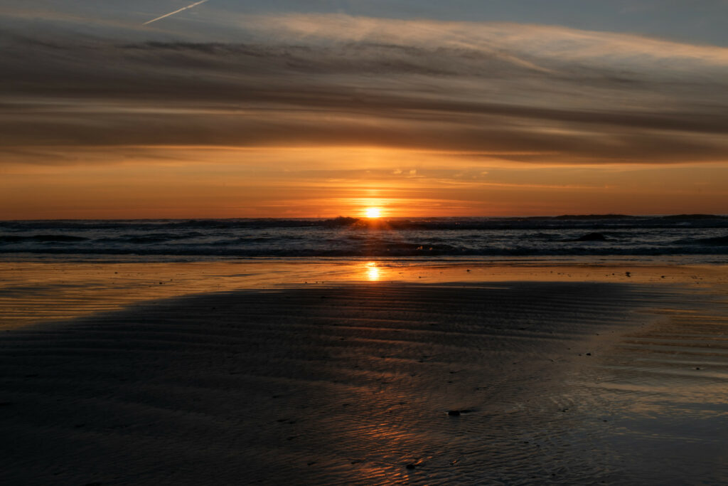 Sunset over sea with glowing sun reflecting on rippled wet sand and soft layered clouds Wallpaper