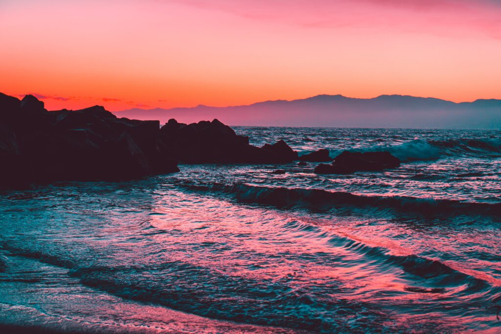 Sunset over sea with pink orange purple sky gentle waves and dark rocks on tranquil shore wallpaper