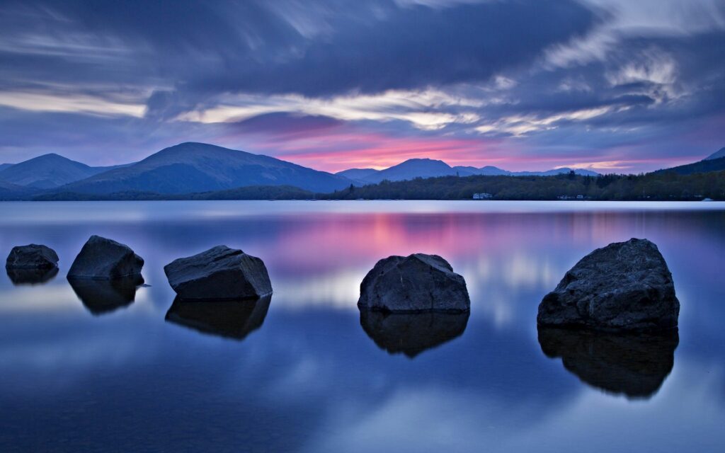 Smooth stones in a tranquil lake at sunset with mountains hills and lush greenery background wallpaper