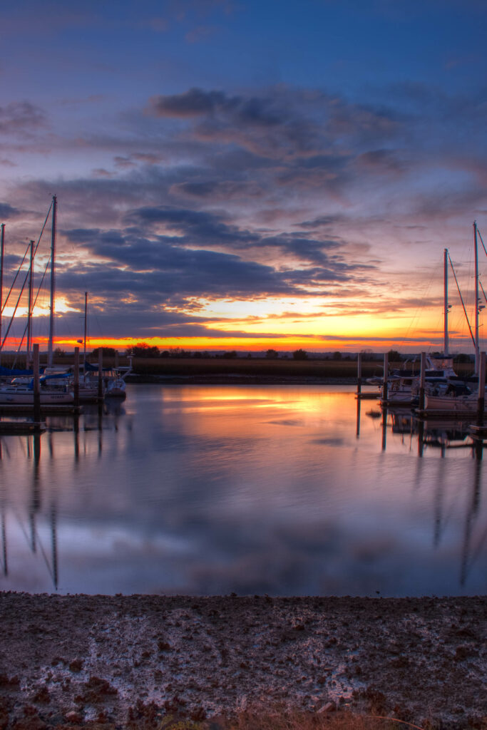 Sunset pier with moored boats reflecting on calm water under warm orange purple sky and clouds Wallpaper