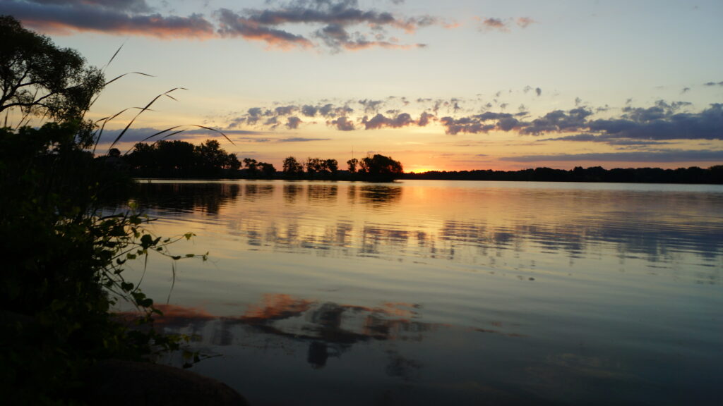 Tranquil river at sunset reflecting warm hues with silhouetted branches and soft clouds background Wallpaper