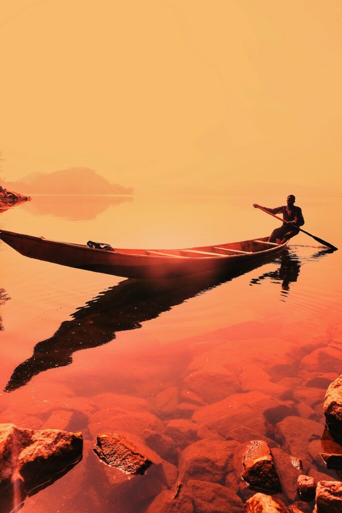 Man rowing wooden boat on calm sea at vivid orange sunset with rocky foreground wallpaper background