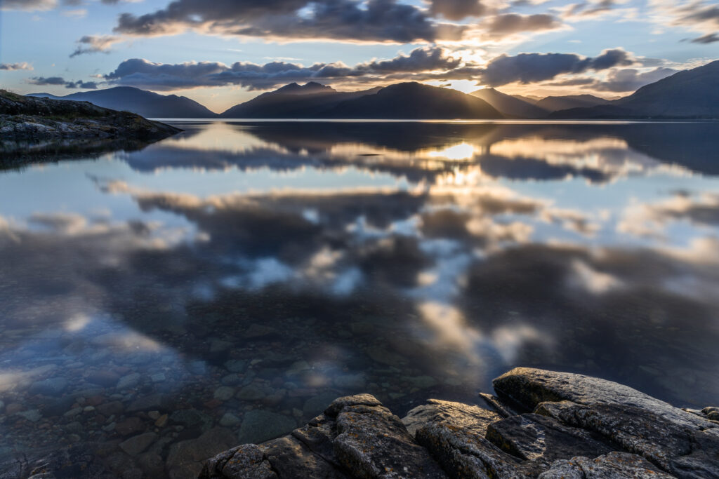 Tranquil sunset sea with clear waters reflecting clouds mountains and smooth stones wallpaper background
