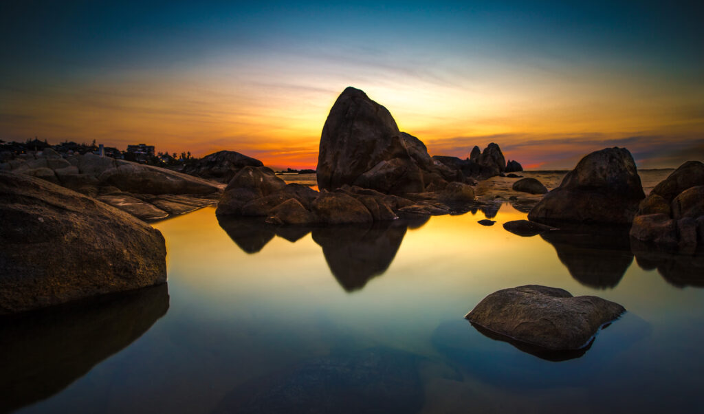 Sunset seascape with large stones reflected in calm water warm orange yellow and blue tones Wallpaper