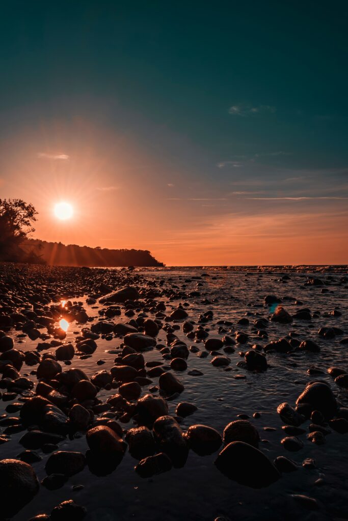 Sunset over tranquil shore with golden light on smooth stones and serene orange and blue hues Wallpaper