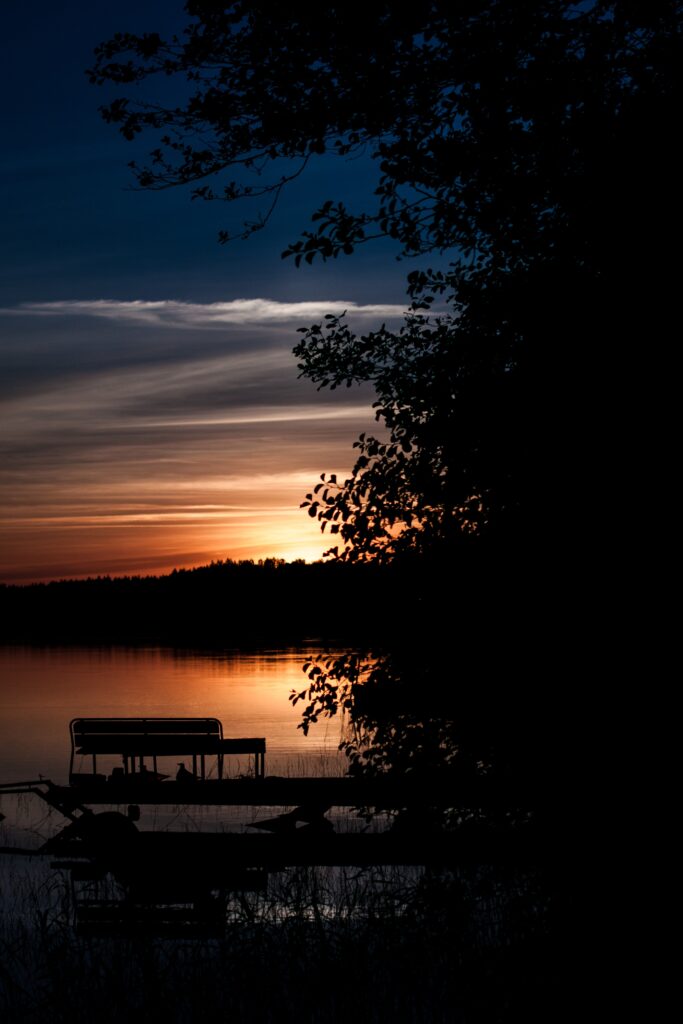 Silhouetted tree and bench by water at vibrant sunset sky with orange and deep blue hues wallpaper background