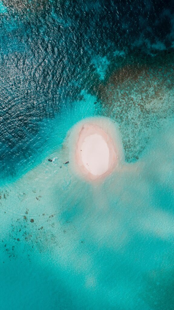 Aerial view of tiny white sand island surrounded by vibrant turquoise and deep blue ocean water Wallpaper