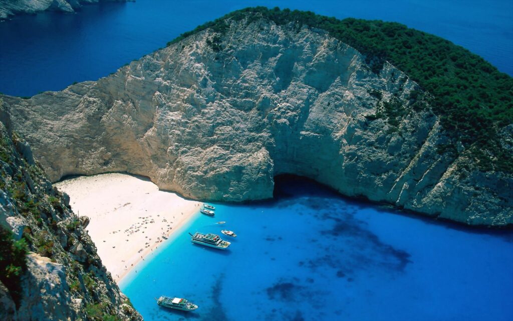 Tranquil coastal scene with turquoise water white sand rocky cliffs and boats under blue sky Wallpaper