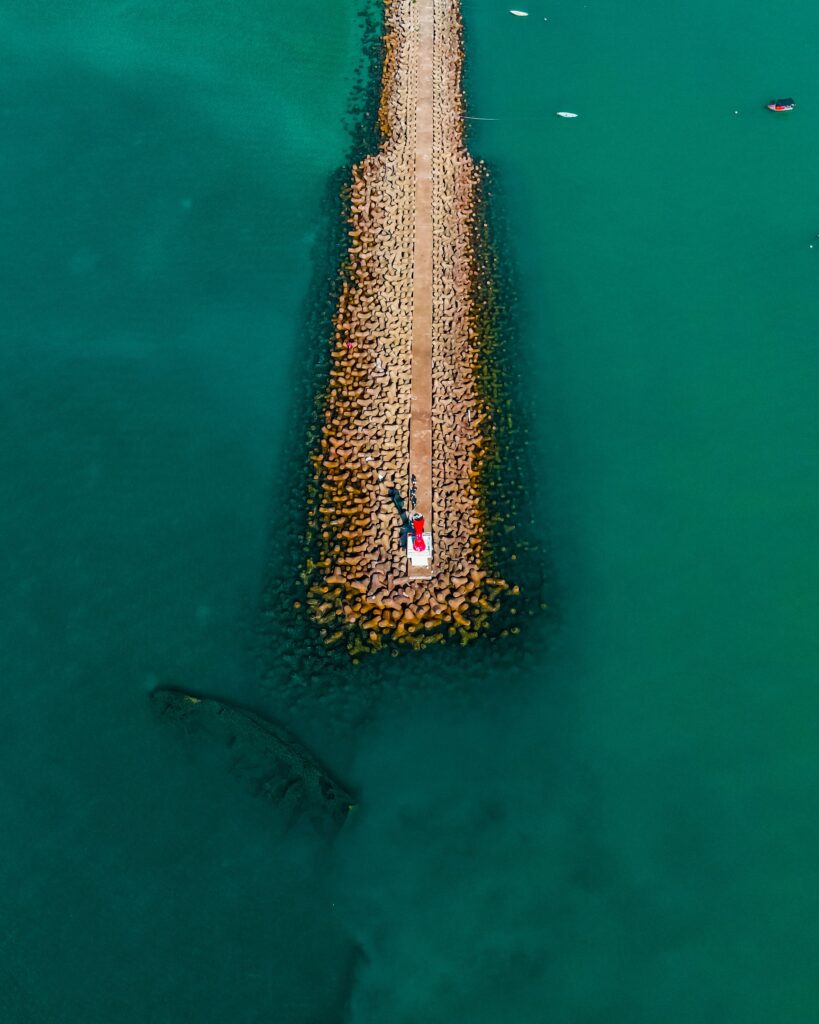 Aerial view of stone pier extending into turquoise water with people and red object center Wallpaper