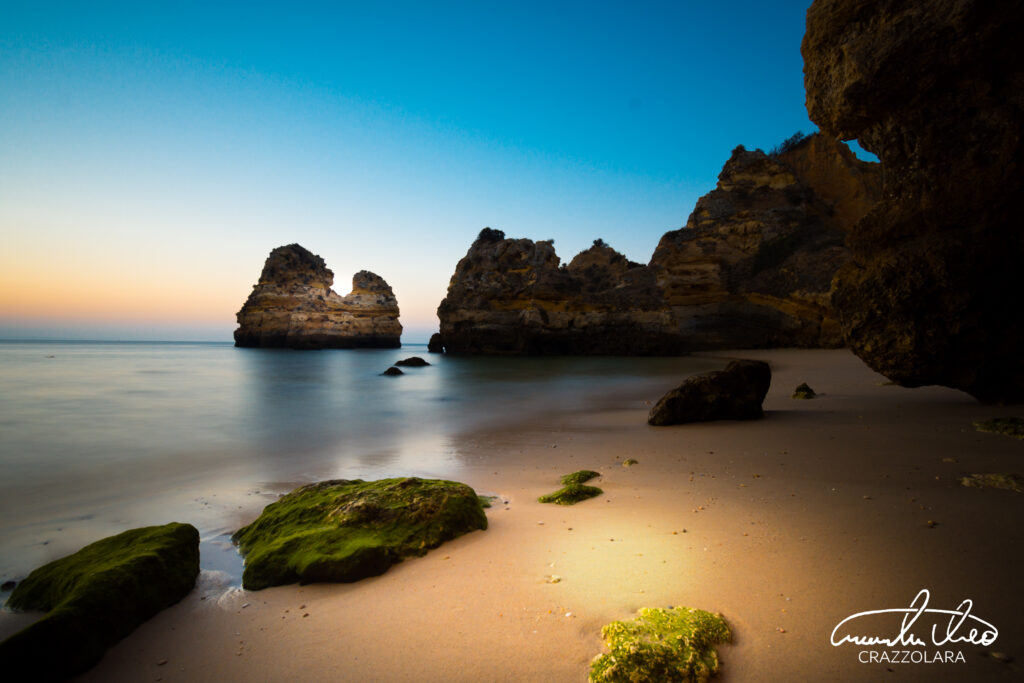 Tranquil twilight beach with dramatic rock formations and mossy stones bathed in soft sunlight Wallpaper