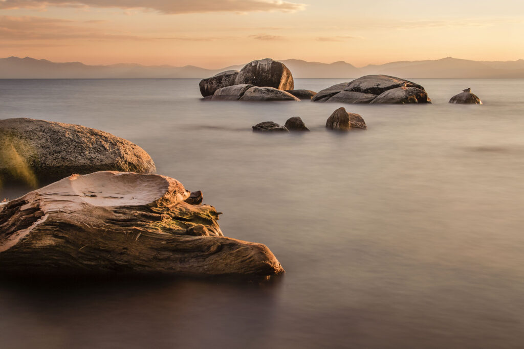 Tranquil twilight sea with large rocks golden peach hues and distant mountains wallpaper background