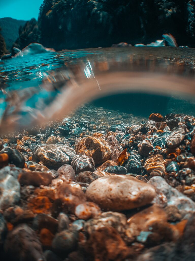 Underwater view of colorful stones in clear water with sunlight and serene natural landscape above Wallpaper