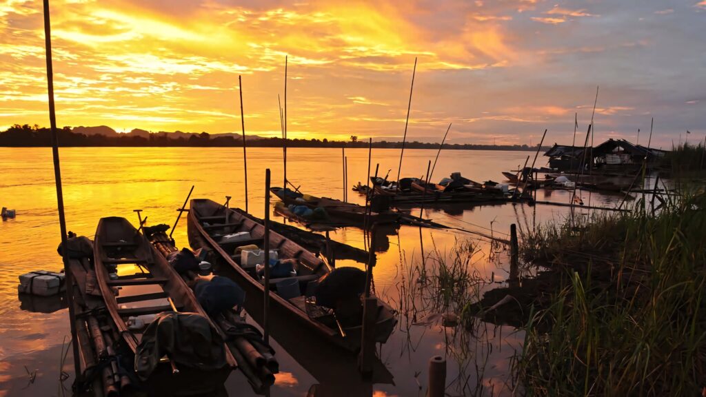 Longboats resting on Vientiane riverside at vibrant sunset reflecting golden calm waters wallpaper