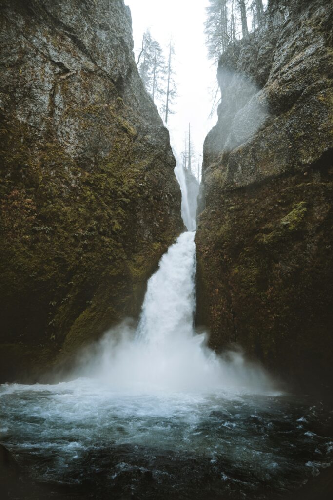 Waterfall cascading between moss-covered rocks mist rising river surrounded by cliffs and trees Wallpaper
