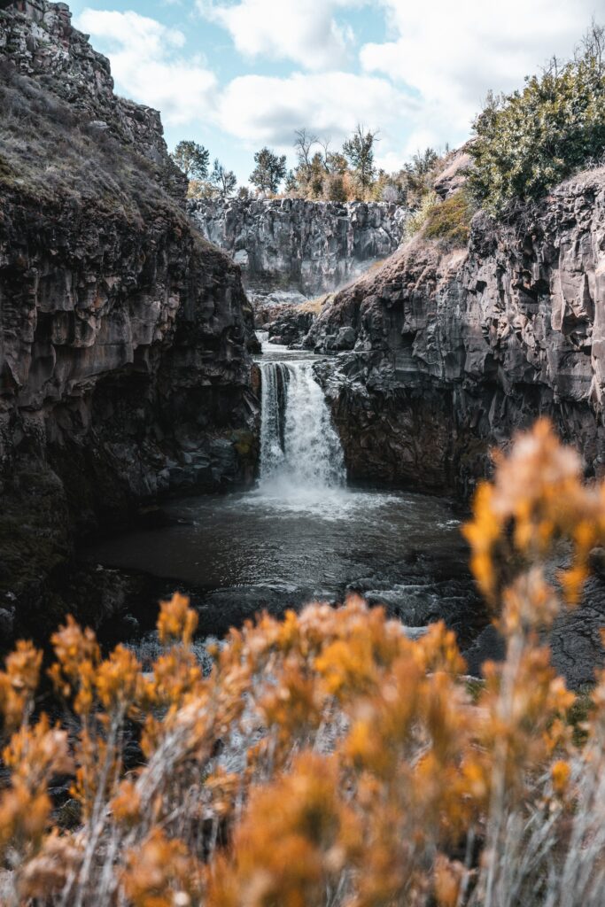 Waterfall cascading through rocky cliffs with autumn leaves and bright sky natural landscape wallpaper