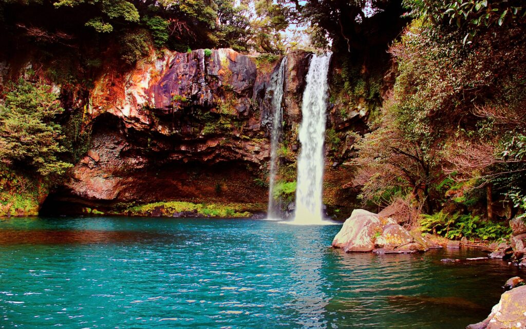 Waterfall cascading into turquoise pool surrounded by lush green foliage and rugged rocks wallpaper
