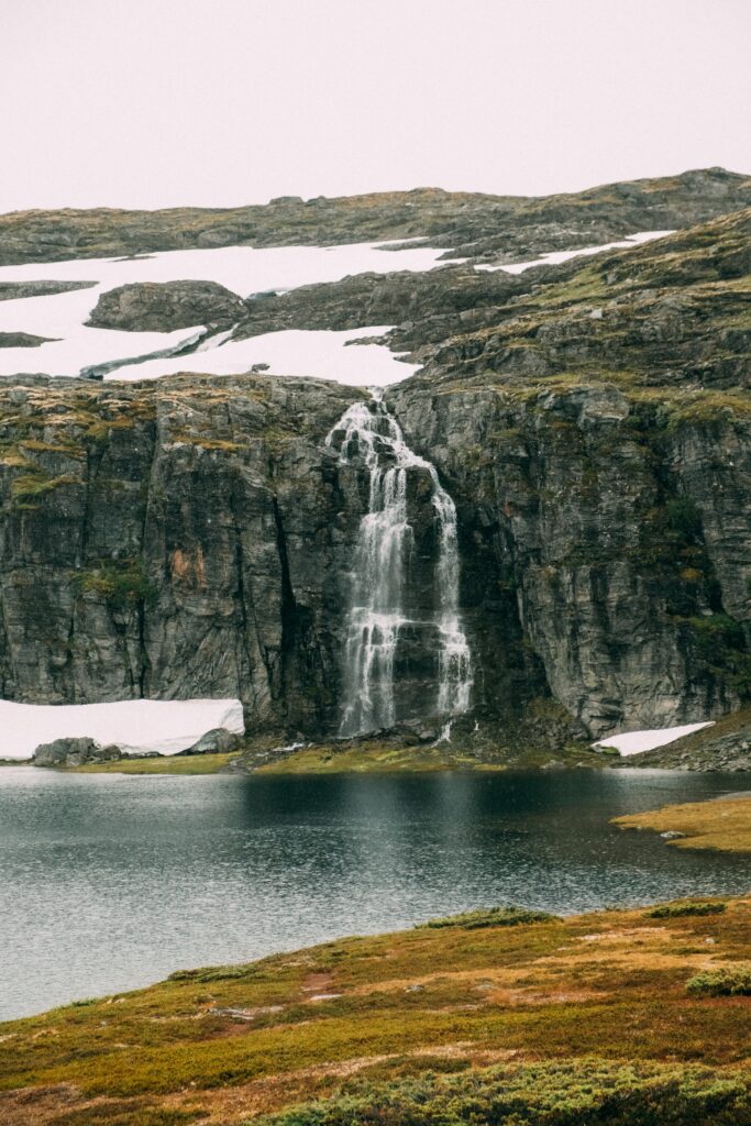 Tranquil waterfall flowing over rugged cliff into serene sea with stones and melting snow background Wallpaper