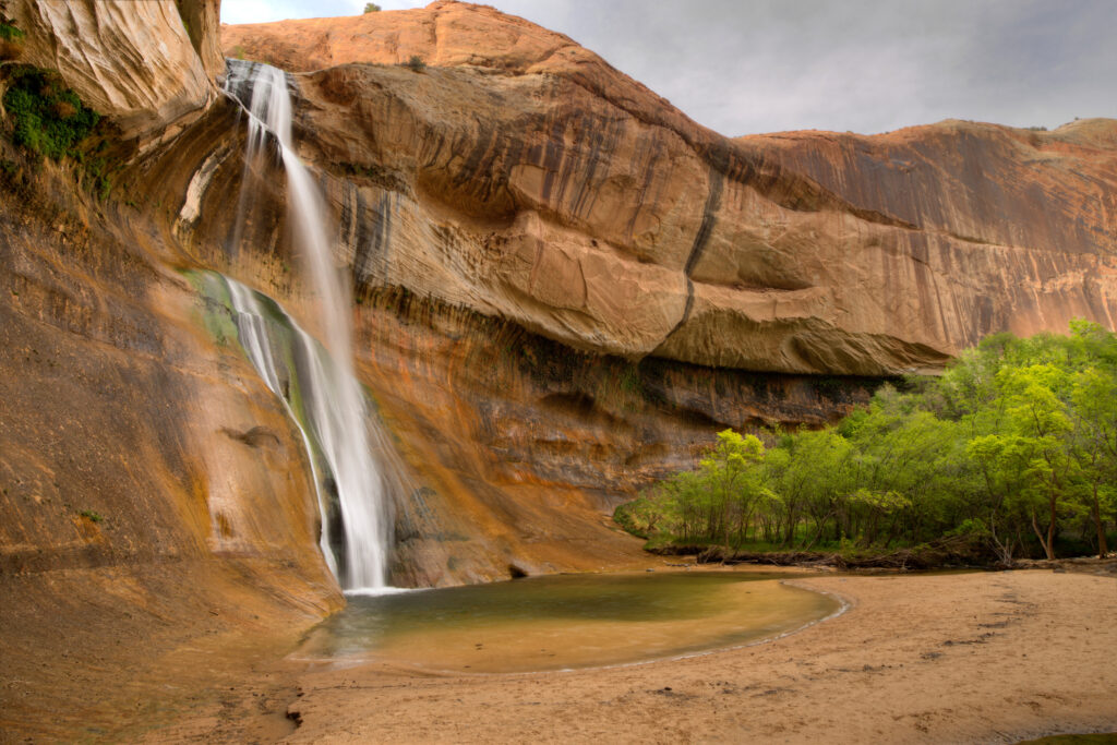 Waterfall cascading down rocky cliff into clear river surrounded by green trees and sandy shore Wallpaper
