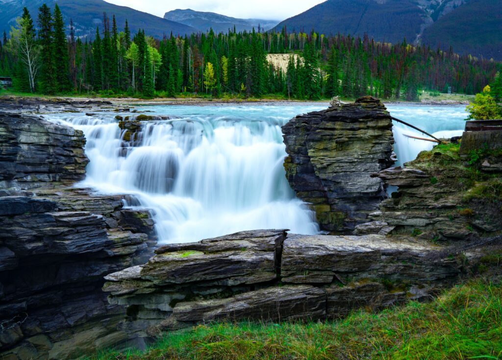 Waterfall cascading over rocky cliffs surrounded by green trees and distant mountain landscape Wallpaper